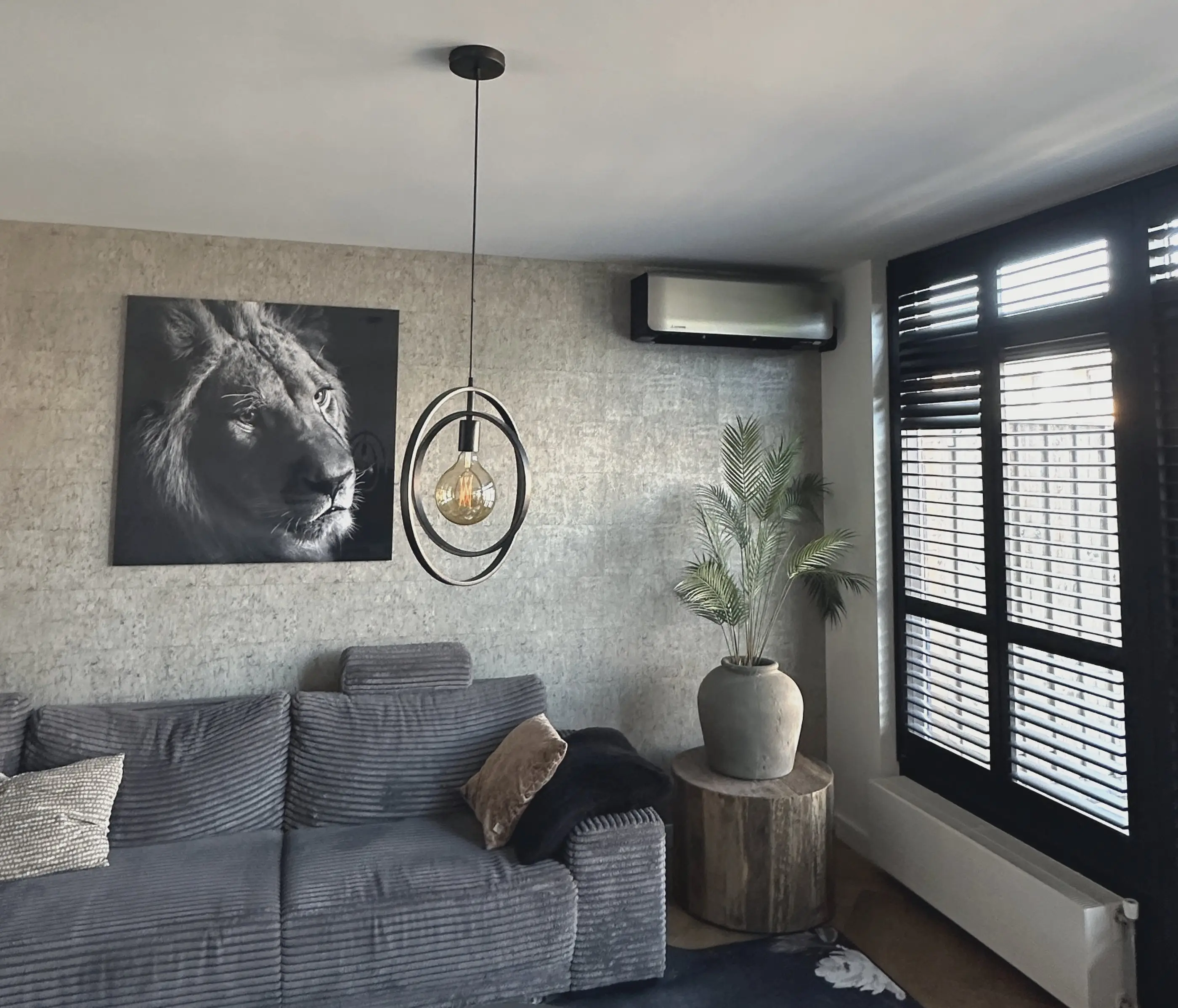 Een woonkamer met een grijze bank, een groot zwart-wit schilderij van een leeuw en hoog aan de wand een strakke binnenunit van de airco.